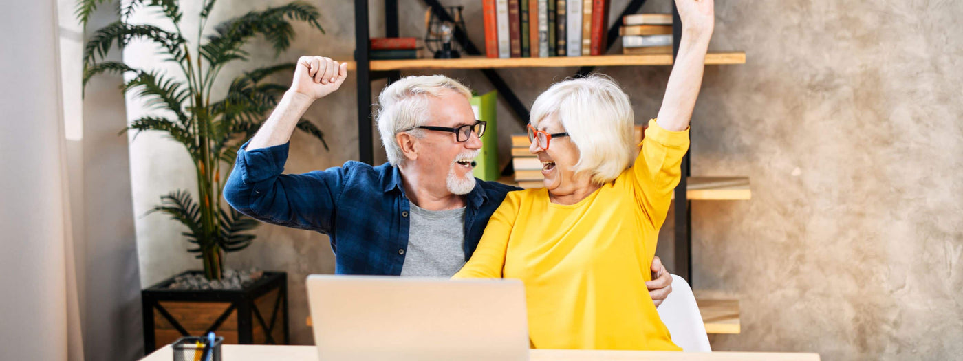 Older couple cheering as they sit at their kitchen table using their laptop